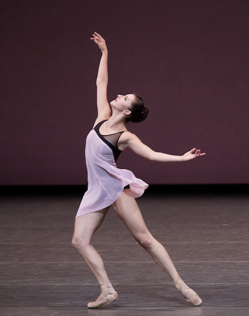 A female dancer in a peach and aubergine filmy costume outstretches her hand overhead and strikes a lunge positionge